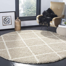 Load image into Gallery viewer, A round shaggy rug in beige and ivory colors, placed on a black floor in a room with a chair, ottoman, and decorative items.