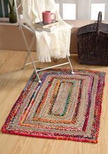 Load image into Gallery viewer, A multicolored braided jute rug with geometric patterns, displayed on a wooden floor next to a chair with a book and a cup, with a wicker basket in the background.