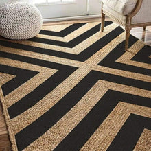 Load image into Gallery viewer, A braided natural black geometric jute area rug with a herringbone pattern, placed on a wooden floor in a home setting.