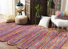 Load image into Gallery viewer, Colorful braided cotton and jute area rug with a vibrant pattern, placed on a wooden floor in a living room setting with various decorative pillows and a traditional wooden cabinet in the background.
